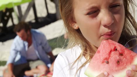 Girl-eating-watermelon-while-father-sitting-in-background-at-beach