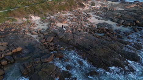 Two-surfers-carrying-surfboards-walking-on-huge-rocks-toward-the-ocean-at-sunset,-dynamic-aerial