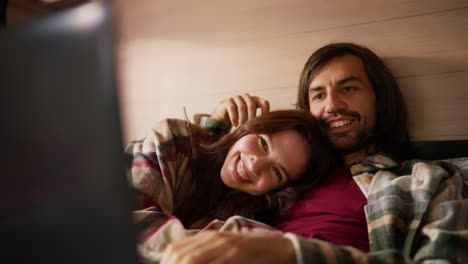 A-happy-brunette-girl-in-a-checkered-shirt-lies-on-the-chest-of-her-brunette-boyfriend-with-stubble-in-a-Green-checkered-shirt-and-they-watch-a-movie-together-using-a-laptop-lying-on-the-sofa-in-a-trailer-in-the-summer-outside-the-city-in-the-camp