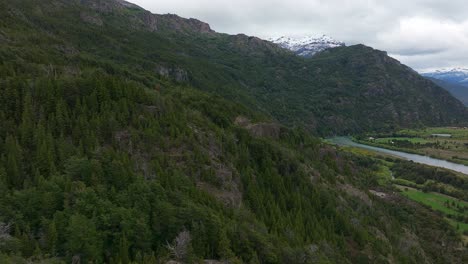 Vuelo-Aéreo-Sobre-La-Ladera-Boscosa-Al-Lado-Del-Río-Futaleufu-Con-Montañas-Cubiertas-De-Nieve-En-El-Fondo