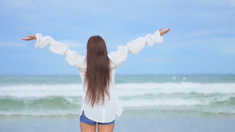 A-young-pretty-woman-walking-along-toward-the-ocean-waves-raises-her-arms-in-joy-as-she-twirls-around-from-the-crashing-waves-to-the-shore