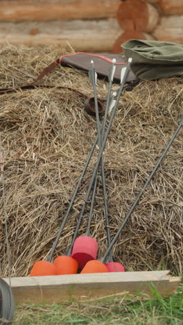 bow and arrow tipped for ignition lie near pile of straw. weapons of medieval warrior and cloak near log cabin in village. historical weapons of ancient heroic campaigns