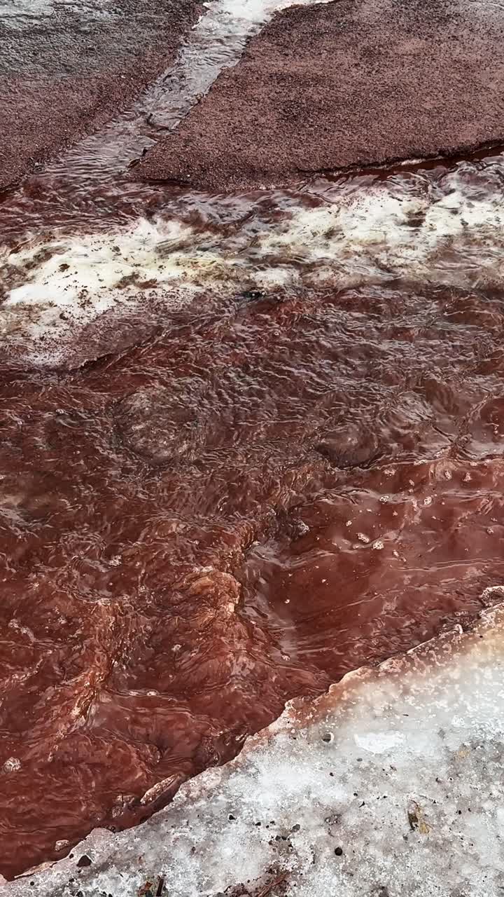 Rapida acqua di disgelo primaverile scorre attraverso un sentiero rosso in disgelo bordato di fanghiglia, trasformando la superficie in un vivido segno a livello del suolo dell'inverno che cede il passo all'inizio della primavera