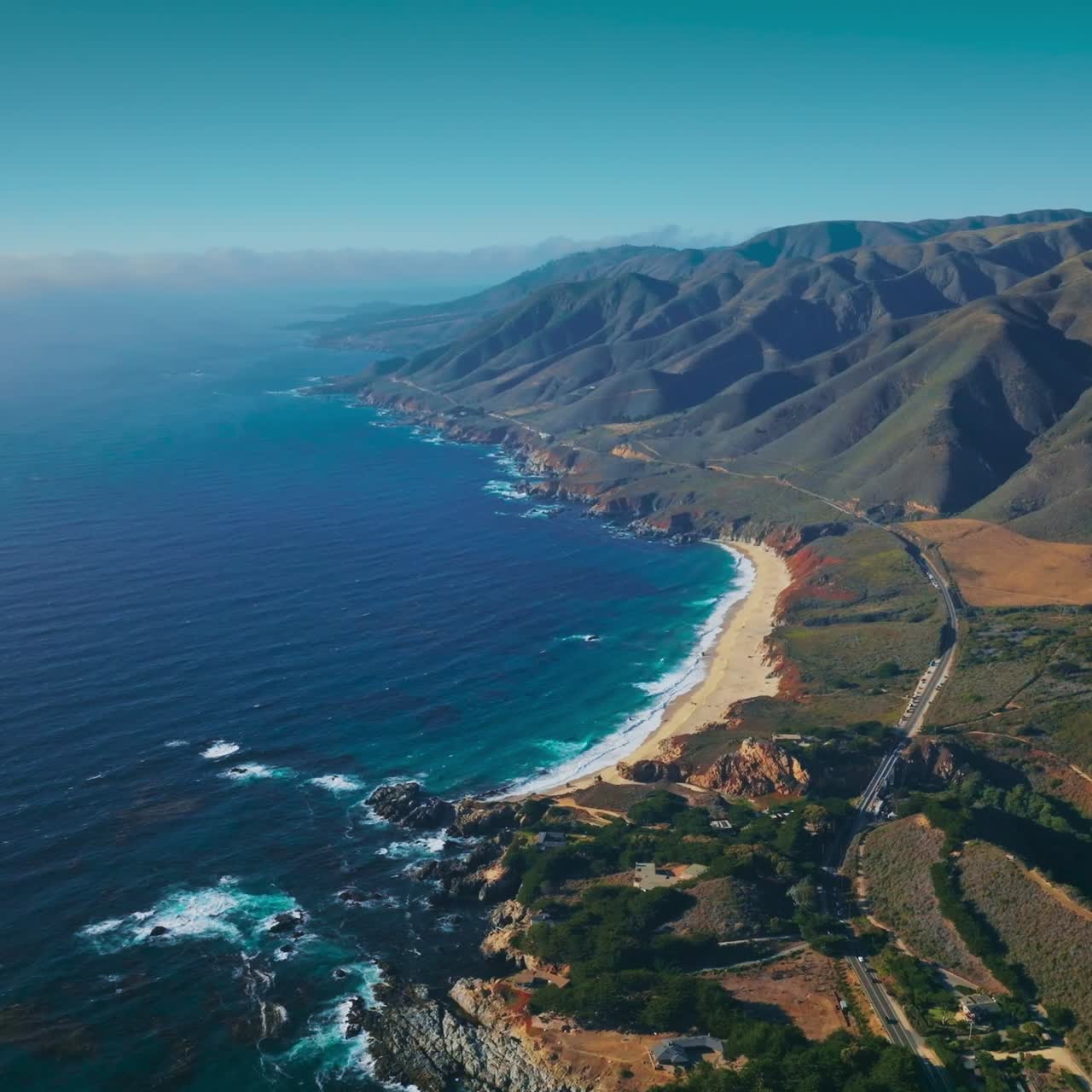 Green inhabited locality among the bare rugged mountains of California. Endless sight of the ocean with hazy horizon at backdrop. Aerial view