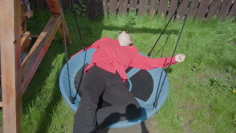 entrepreneur in red shirt lays comfortably on round blue swing, adjusting posture and soaking in warm sun, scene set in vibrant green backyard with wooden swing frame