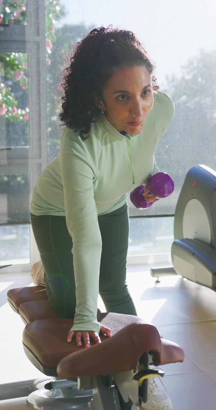 Vertical video: African American woman bracing left hand, doing one-arm row on bench with dumbbell