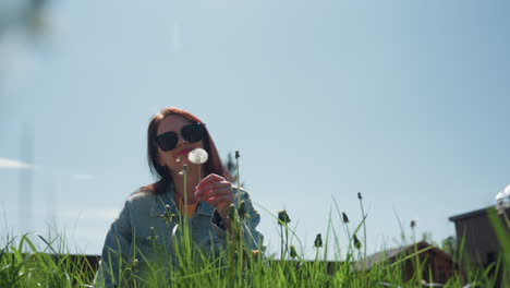 Woman wearing denim jacket and sunglasses blows dandelion while sitting in tall green grass, enjoying bright sunny weather with clear blue sky