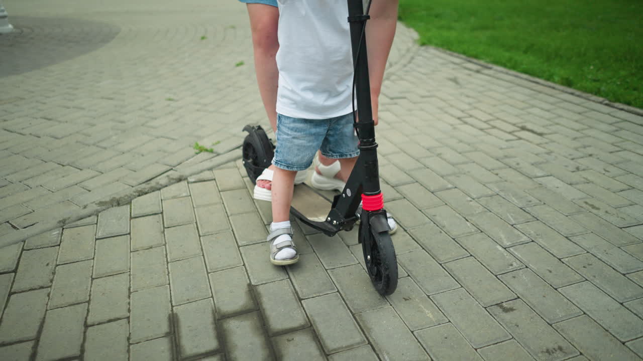 A close-up leg view of a child and woman standing on a scooter along an interlocked path outdoors