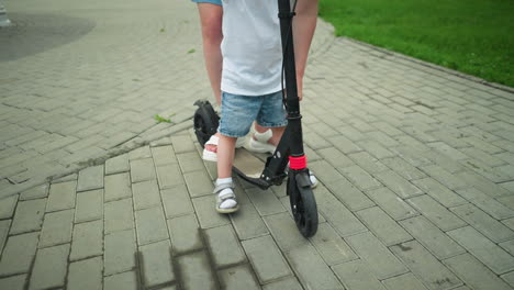 a close-up leg view of a child and woman standing on a scooter along an interlocked path outdoors