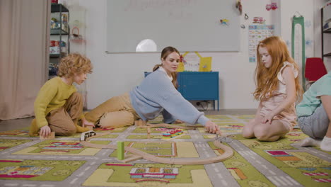 little blond boy and redhead little girl playing with their teacher with wooden cars on the carpet in classroom in a montessori school