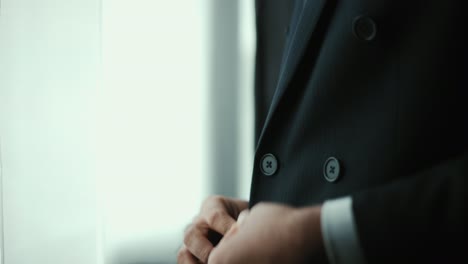 confident businessman buttoning or adjust classic blue suit near window in hotel room at the morning. handsome man wearing a nice suit on wedding day.