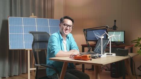 hombre asiático mira la turbina eólica y sonríe a la cámara mientras trabaja con una computadora portátil junto a la célula solar en la oficina