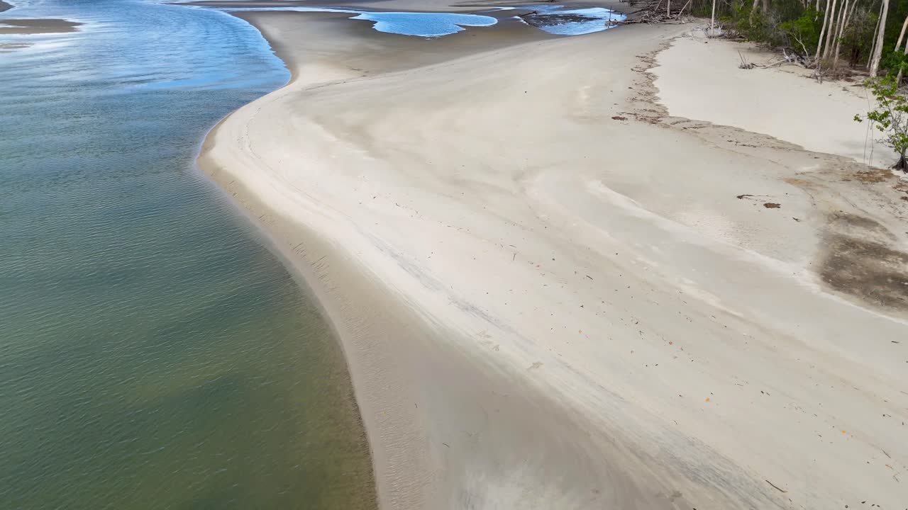 Drone glides above tranquil river meeting sandy shore, bordered by lush rainforest in daylight