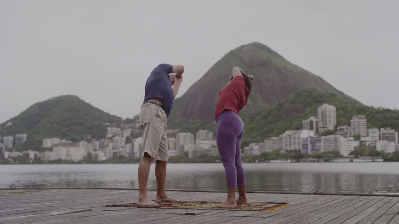 Couple practicing yoga or stretching outdoors by a lake with mountains and a city skyline in the background