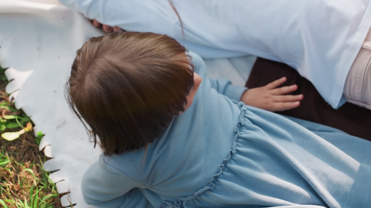 Aerial view of little girl lying faced up next to sister on soft blanket outdoors, relaxing thoughtfully under daylight, showing warmth, sibling bond, calmness, and peaceful childhood atmosphere