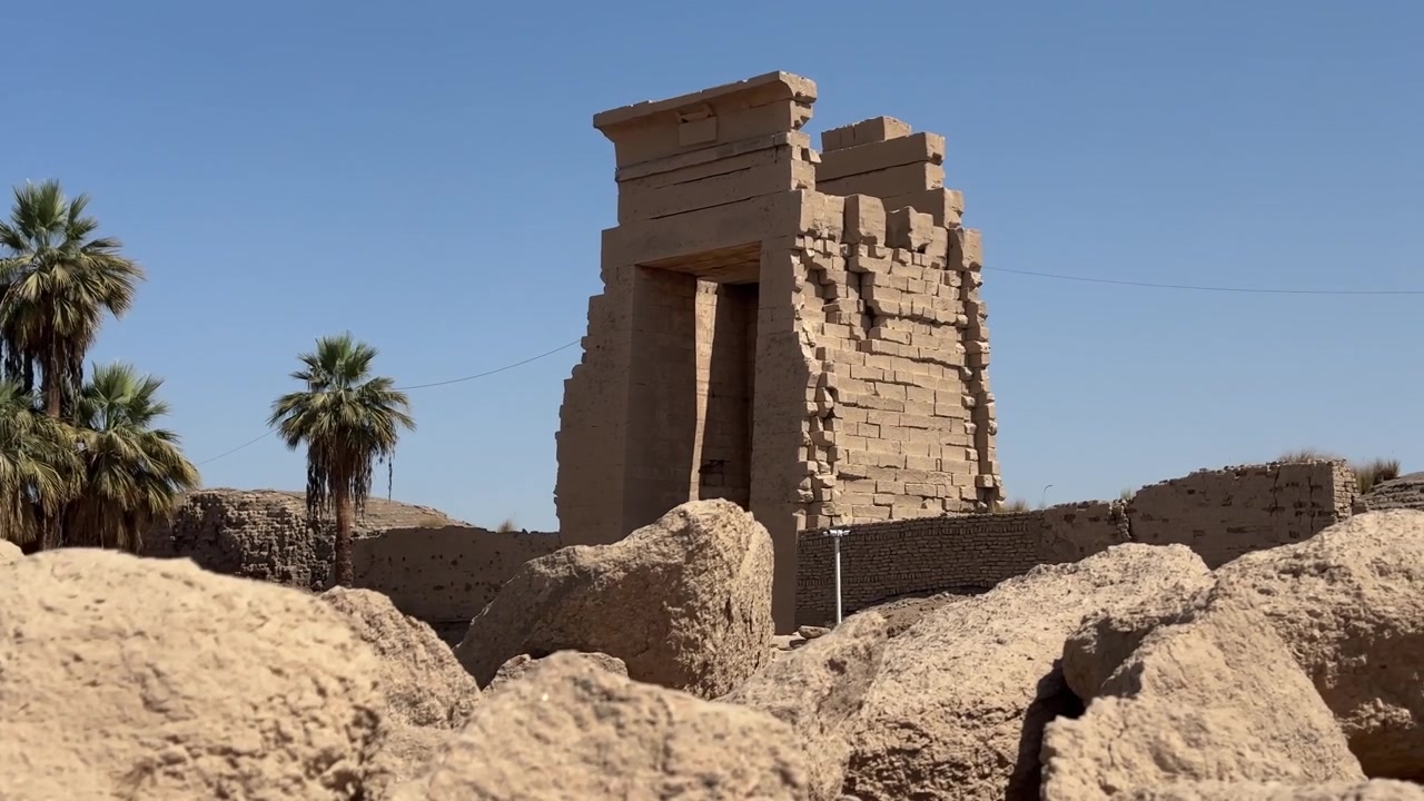 Sebuah bidikan sinematik statis dari gerbang pilon batu monumental di kompleks Kuil Karnak di Luxor, menampilkan arsitektur Mesir kuno dan pohon palem di bawah langit cerah.