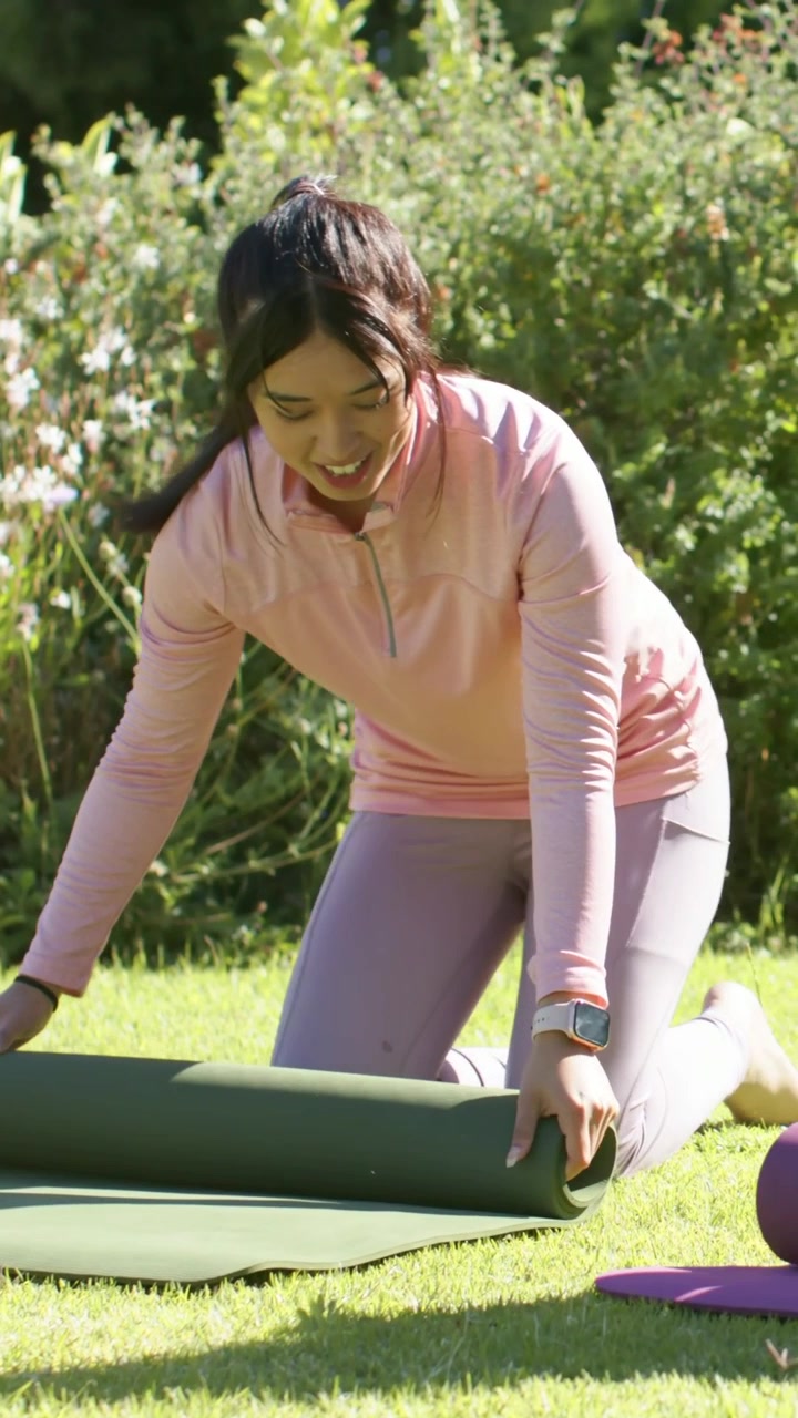 Video vertical: Mujer sujetando una esterilla verde, desenrollándola en un parque, preparándose para el entrenamiento, con top rosa y un accesorio morado