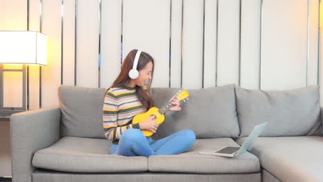 a pretty young woman, wearing headphones, sits on a comfortable sofa learning to play the ukulele from an online source