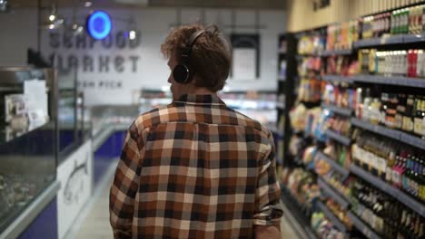 stylish caucasian guy with headphones walks through goods section of the store, wearing plaid shirt - listening to the good music.. following back view shot. slow motion