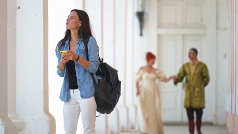 young woman tourist with a backpack looks at her phone wants to take pictures of an old castle. couple wearing historic costumes blurred walking up to a girl. history repeats, past meets present.