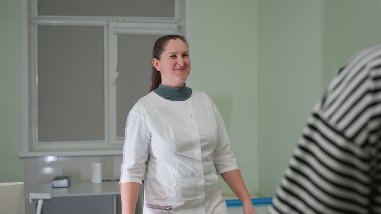 Medical doctor stands from desk to warmly welcome patient into clinic, directing her with friendly smile to examination bed while maintaining professional composure in well-lit clinical environment