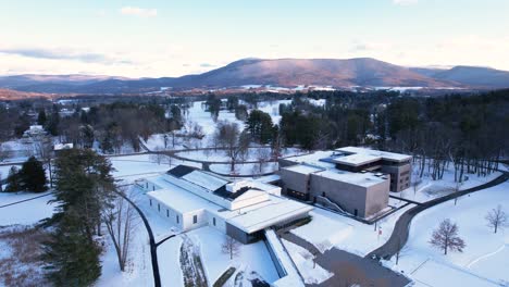 low altitude flyover of museum of arts among snow blanketed fields, frozen ponds, and evergreen trees, rolling hills in the background on a scenic winter landscape