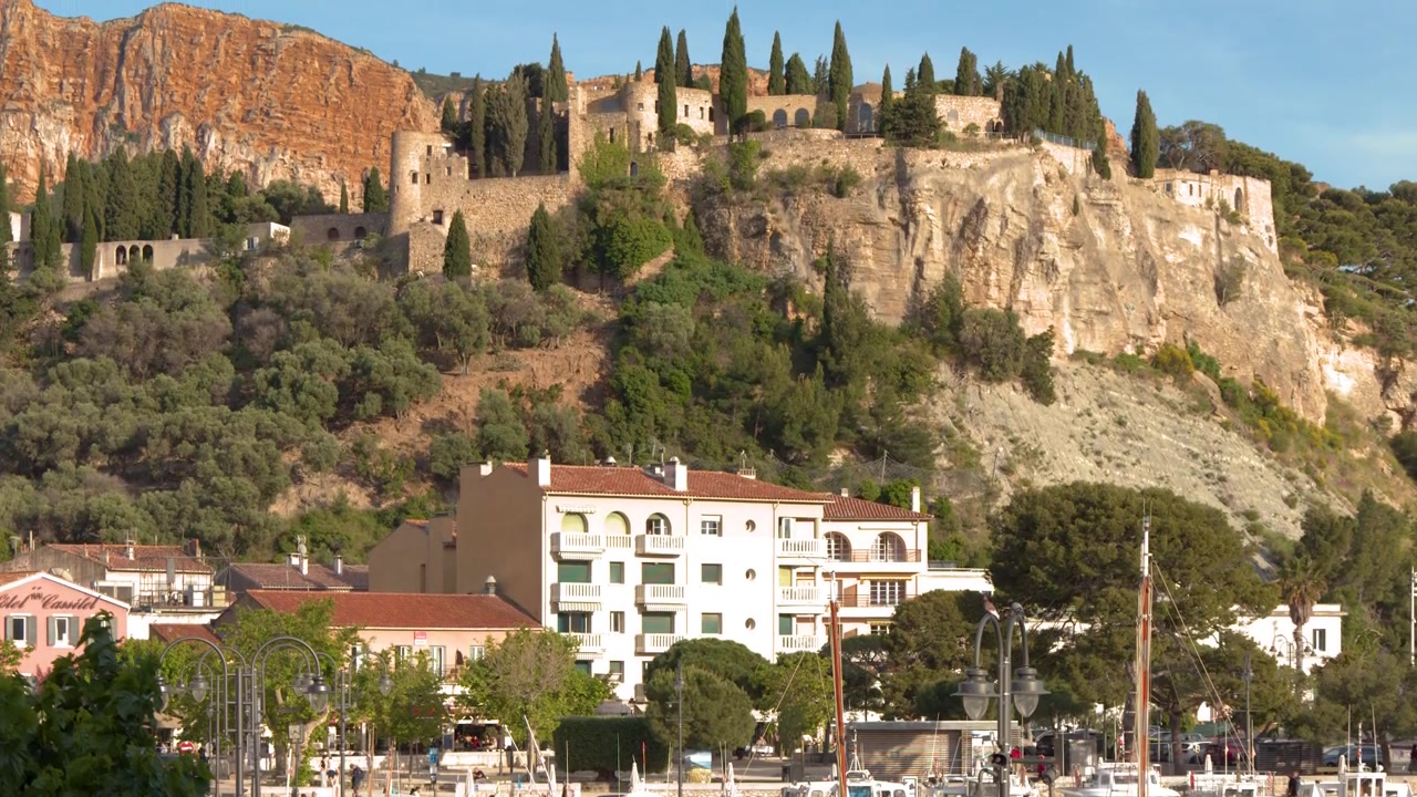 Inquadratura ampia fissa di un porto soleggiato con barche ormeggiate sotto scogliere calcaree ed edifici