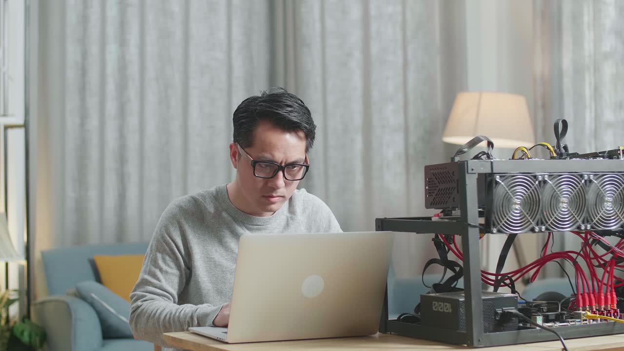 안경을 쓴 아시아인 남자가 암호화폐를 채굴하기 위해 채굴 기계를 가진 노트북을 사용하고 있습니다.