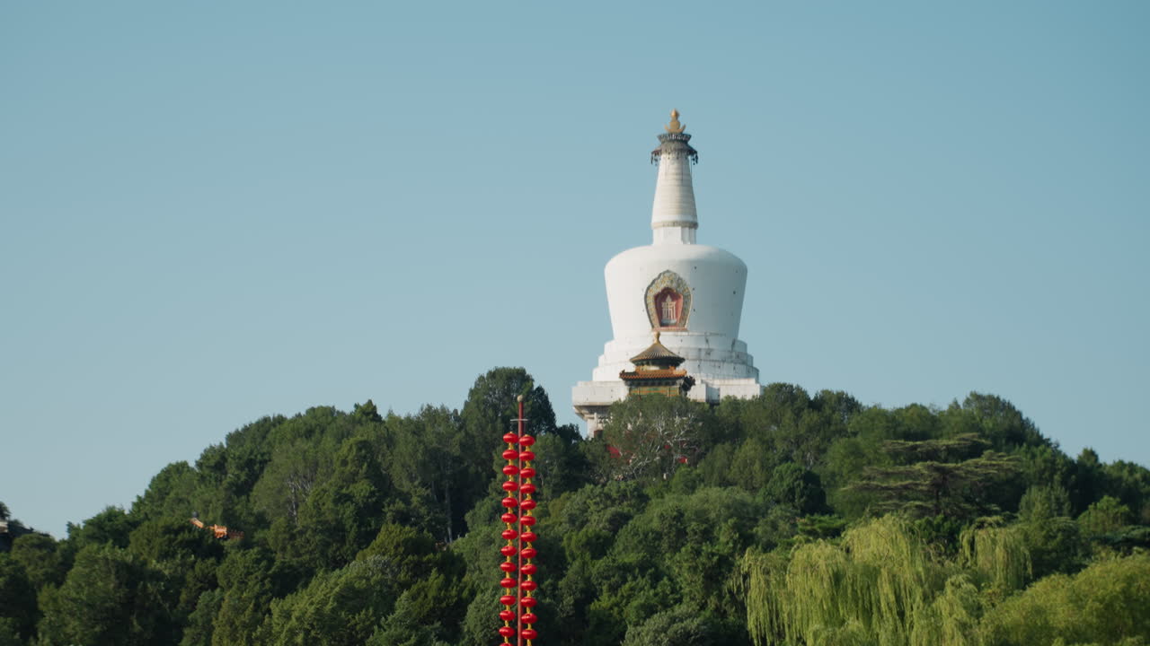 Estupa blanca en lo alto de una colina verde en Beijing. Una vista serena al mediodía que muestra una estupa budista alzándose sobre árboles frondosos bajo un cielo azul claro. Un poste ornamental con linternas rojas en primer plano y una mampostería detallada. Una escena de gran calma.