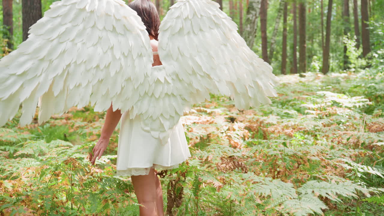 Back view of angel girl in white dress walking gracefully along forest path holding wooden staff among green leaves and golden ferns under warm sunlight