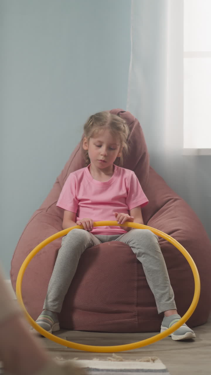 Unhappy little girl with hula hoop sits in bean chair while mother works on laptop at home slow motion. Preschooler child bored to be quite. Remote work