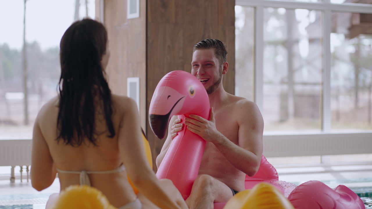 una pareja adulta está descansando en un parque acuático. un hombre y una mujer están sentados en círculos de natación inflables bromeando y riendo.