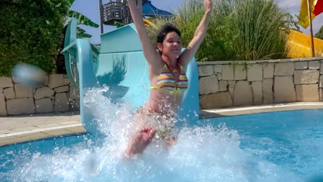 descenso del tobogán acuático en un parque acuático de vacaciones. cámara lenta en un tobogán acuático vacaciones familiares, una mujer en bikini desciende del tobogán en una piscina de agua azul salpicando gotas de agua.