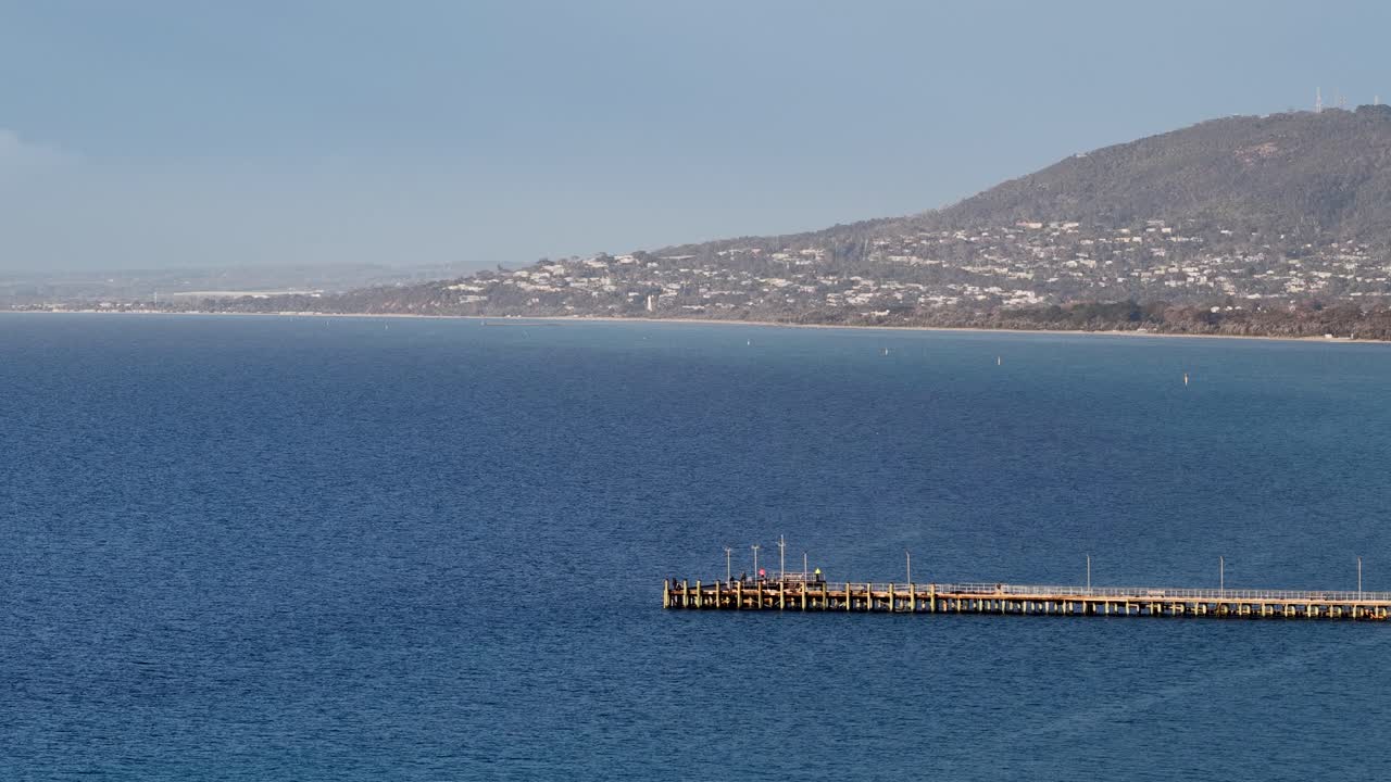 Wide daytime pan of wooden pier over blue bay, revealing coastline, distant hills, clear sky