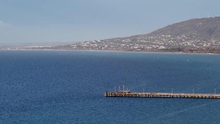 Ampia panoramica diurna di un molo di legno su una baia blu, che rivela la costa, le colline in lontananza e il cielo sereno