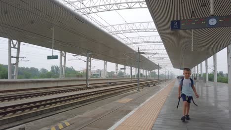 little caucasian boy with a small backpack walking on an empty platform while waiting for the arrival of the super fast chinese high speed bullet train