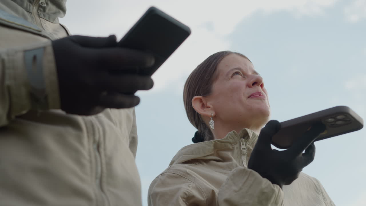 close up adult wearing gloves holding smartphone while companion looks up contemplatively against bright sky with soft background blur and warm sunlight reflection in lush green field