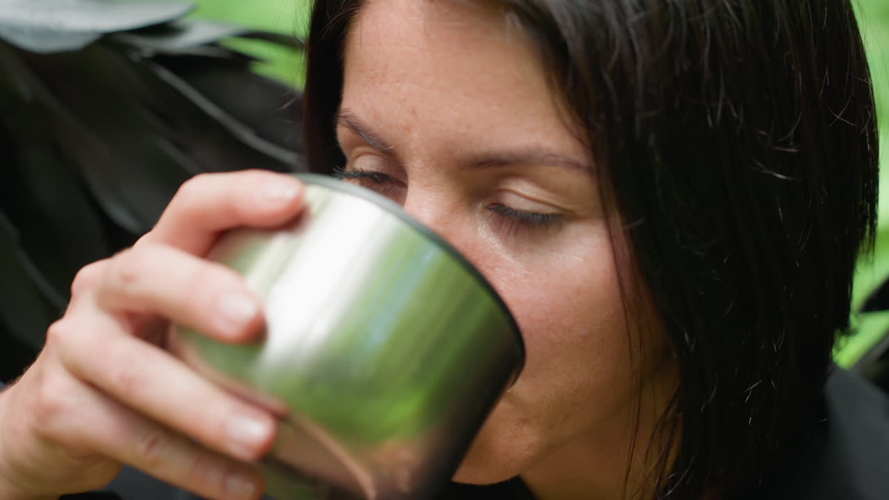 Close up of fallen goddess in dark costume holding stainless flask cover and sipping drink in forest setting, soft light reflecting on metal surface creating serene mood