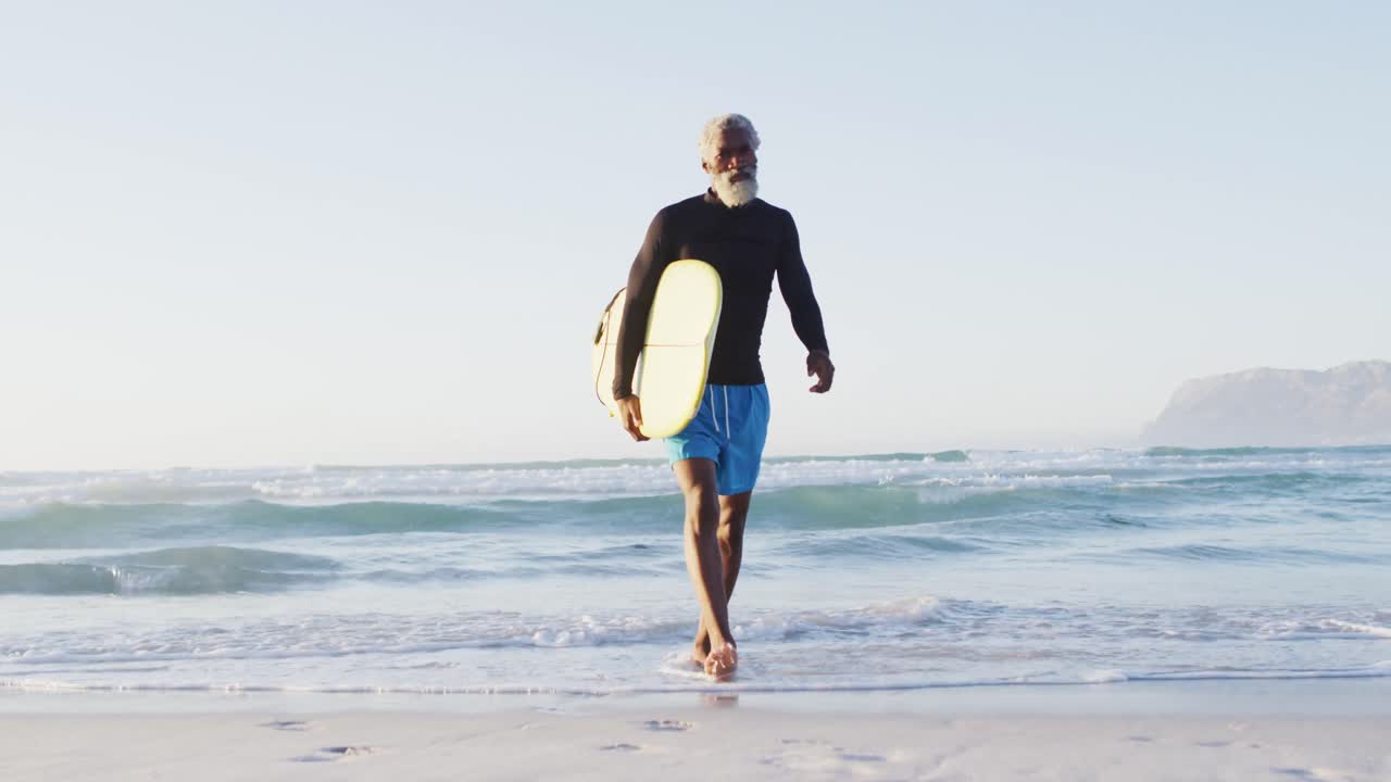 hombre afroamericano mayor caminando con una tabla de surf en una playa soleada