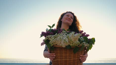 mujer adulta linda con cabello rizado con vestido con canasta retro fuul de lila de pie cerca del mar al amanecer o al atardecer. tiempo de viaje de vacaciones. escena romántica. estilo de vida femenino. 4k