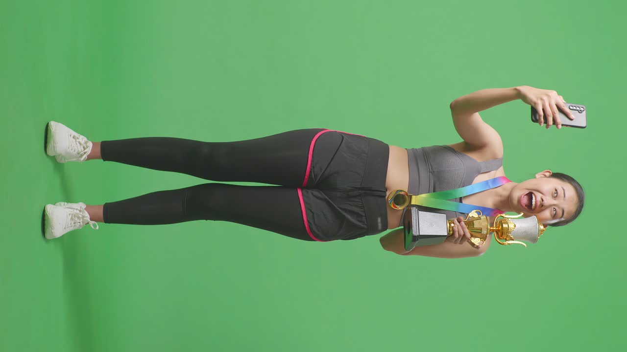 골드 트로피를 들고 스마트폰으로 사진을 찍는 아시아 여성의 몸 전체가 스튜디오의 녹색 화면 배경에서 첫 번째 우승자로