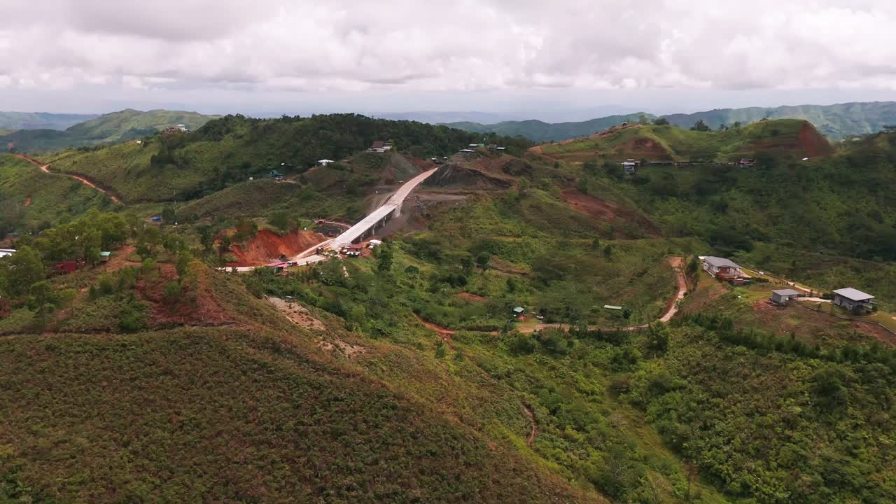 aerial drone shot of a modern highway bridge under construction, cutting through steep, rugged, and densely vegetated hills and rural terrain. Represents infrastructure development in remote areas