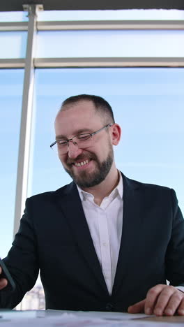 happy businessman in office on window background. portrait of successful entrepreneur sitting at table and reading phone message. vertical video