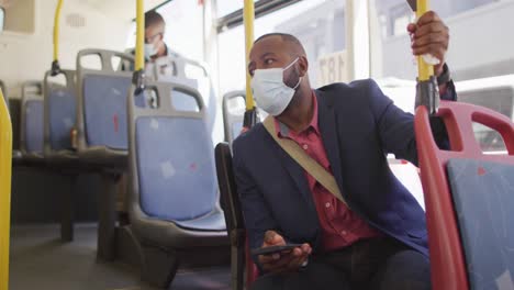 african american man in city wearing face mask, using smartphone in bus