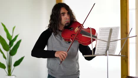 hispanic male musician with long brown hair sternly playing songs from sheet music on red viola in white studio