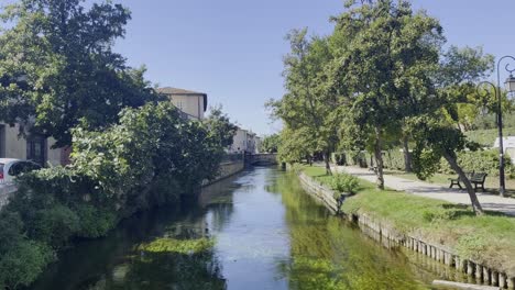 Kleiner,-Breiter-Fluss-In-Frankreich-In-Einer-Kleinen-Stadt-An-Einem-Park-Bei-Schönem-Sonnigen-Wetter-Mit-Kleinen-Steinhäusern-Und-Viel-Natur-Und-Einer-Brücke