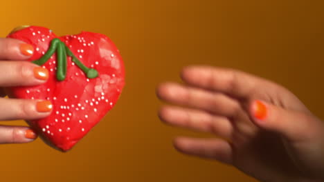 two hands with orange nail polish exchange a heart-shaped red donut decorated with white sprinkles and green icing stems, set against a warm orange background in a playful and sweet gesture.