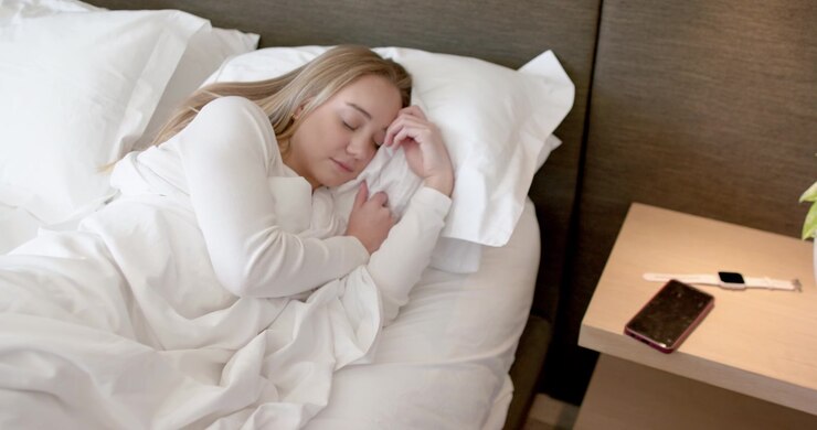 Blød belysning der får en kvinde til at sove, mens hun holder et tæppe i sengen, telefon, ur og plante på natbordet