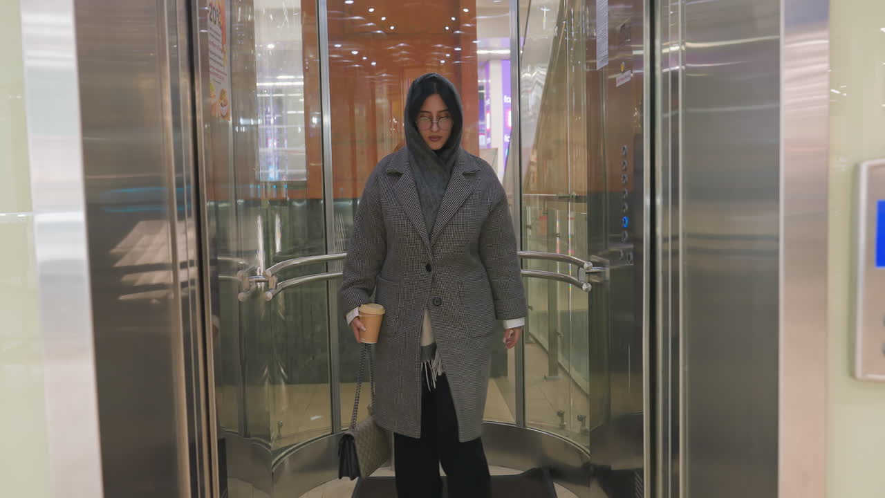 fashionable woman in oversized coat with glasses holding takeaway coffee and handbag steps out elevator into modern building with transparent walls and bright interior reflections in background