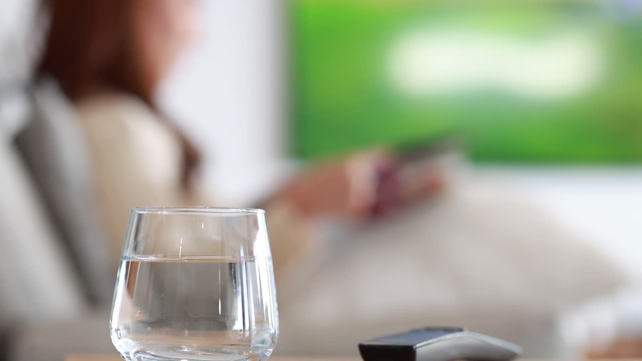 A young Asian woman sits on a modern sofa, holding a remote and watching television. A glass of water and remote rest on a table in soft, natural daylight